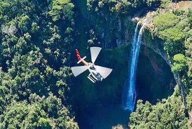Red-and-white helicopter near a tall waterfall descending into a densely forested gorge in Mauritius.
