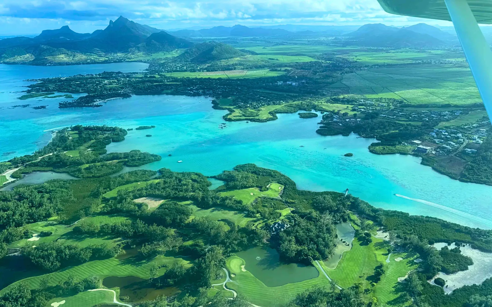 View from a seaplane over Mauritius’s eastern lagoon, with green islands, golf fairways, coastal settlements and mountains in the distance.