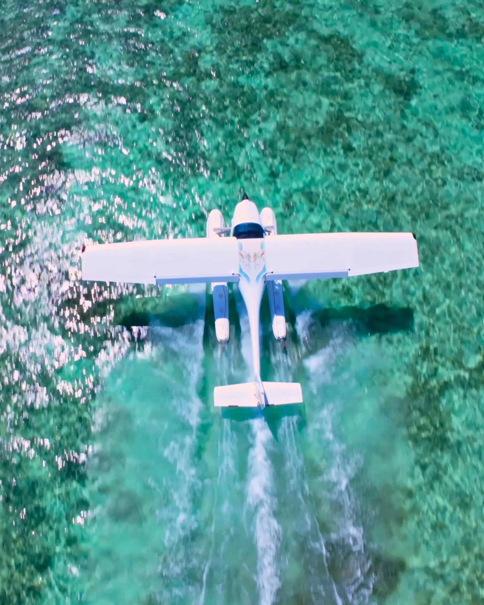 Overhead view of a white seaplane on the clear turquoise lagoon in Mauritius, with its floats and wake visible beneath the wings.